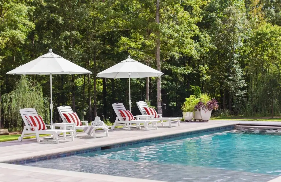 Resort-inspired pool deck featuring white Nautical chaise lounges with striped cushions and large umbrellas beside a swimming pool.